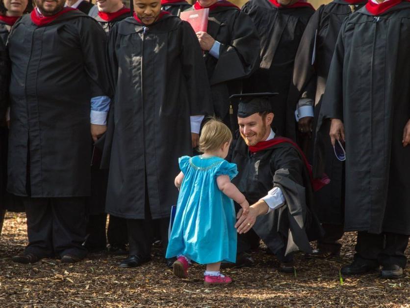 A graduate kneels down to hug his young daughter.